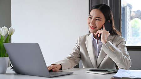 Millennial woman in formal suit checking information on laptop and talking on mobile phone with her...
