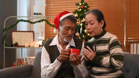 Happy senior couple sitting on couch next to a decorated Christmas tree and enjoying drinking hot...