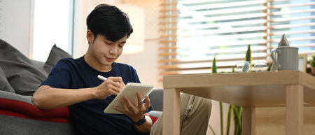 Handsome man freelancer sitting front of laptop in cozy living room and making notes on notebook.の写真素材