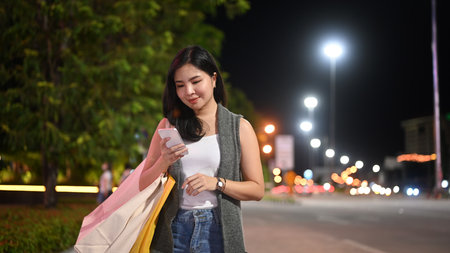 Stylish woman typing text message on her mobile phone while walking through night city street with blurred lights background.の写真素材