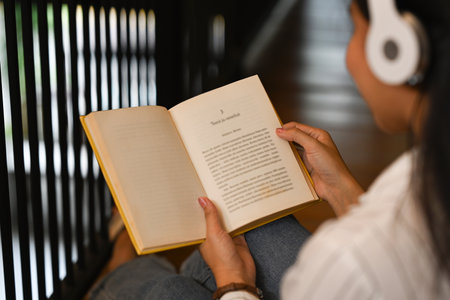 Over shoulder view of young woman wearing headphone and reading book in library. People, knowledge and education concept.の写真素材