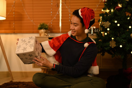 Smiling man in Santa hat opening Christmas gifts and sitting in decorated room for celebrating New...