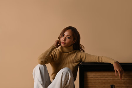 Portrait of fashionable woman wearing trendy sweater sitting near vintage radio, studio shot on beige background.の写真素材