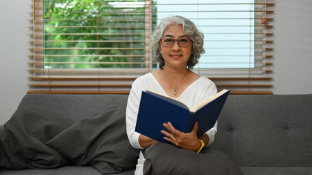 Happy mature woman resting on couch and reading interesting book, enjoying leisure weekend time at...