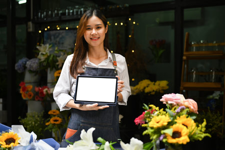 Confident young female florist holding digital tablet standing front of her floral shop. Blank screen for your advertise text.の写真素材
