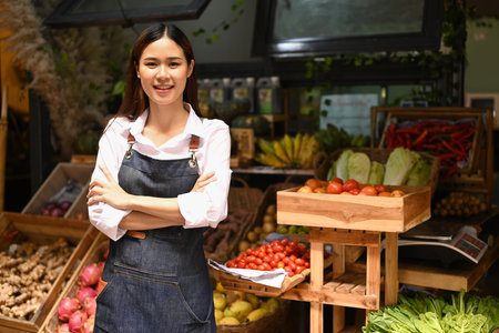 Portrait of friendly woman organic vegetable stall owner in apron standing with crossed arms and...