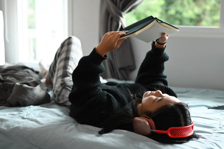 Carefree teenage girl laying on bed in a cozy bedroom and reading book. Leisure and people concept.の写真素材