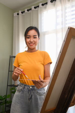 Image of female painter holding paintbrush and palette standing in front of canvas and smiling to camera.の写真素材