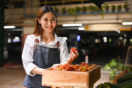 Smiling asian woman vegetable stall owner arranging tomatoes in on store shelves. Farmers market concept.の写真素材
