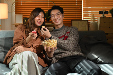 Joyful young couple eating popcorn and watching movie, spending leisure time in cozy winter or...