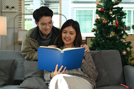 Romantic young couple sitting on couch and reading book, spending leisure time in cozy winter or autumn weekend.の写真素材