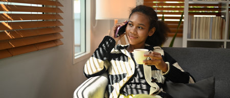 Happy young woman having pleasant phone conversation and drinking hot coffee on couch at home.