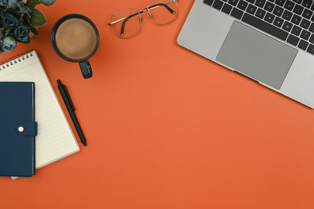 Laptop, cup of coffee, notepad, glasses and flower pot on orange background. Flat lay, Top view with copy space.の写真素材