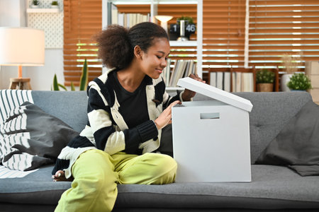 Happy African woman sitting on sofa at new apartment and unpack box with belongings at moving...