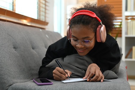 Young African woman listening to music on headphone and doing homework on couch.