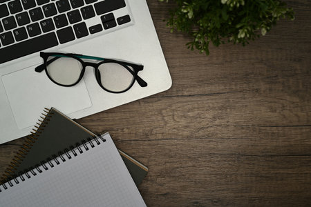Top view of laptop, eyeglasses, notepad and houseplant on wooden office desk. Copy space for text.