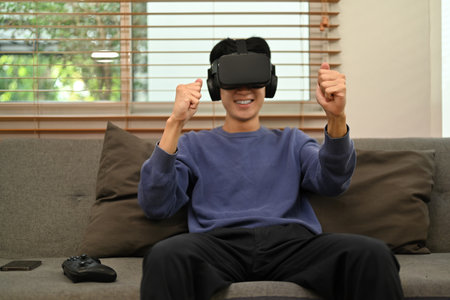 Joyful asian man experiencing opportunities of gaming in virtual reality headset while sitting ouch...
