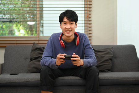 Smiling young asian man playing video game with joystick while sitting on couch at cozy home.