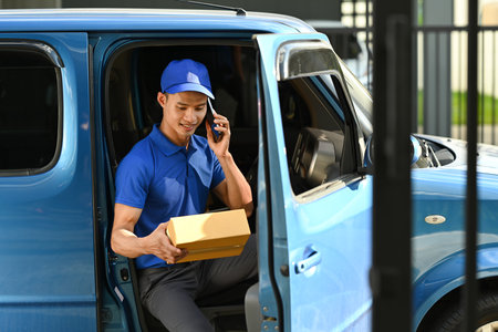 Smiling asian male courier talking on mobile phone while sitting while sitting van. Delivery service and shipping concept.の写真素材