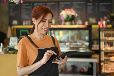Satisfied female entrepreneur wearing apron standing in front of counter of modern cafe using...