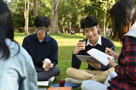 Group of university students talking with and reading book while sitting together on green grass in campus.の写真素材