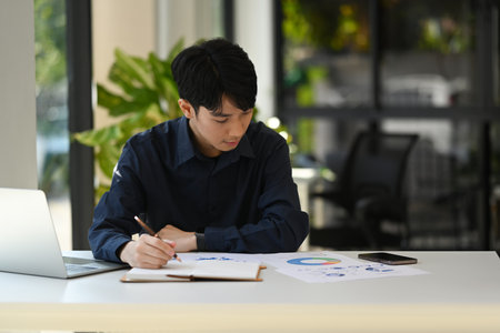 Focused asian male economist using laptop and checking statistic document at office desk.の写真素材