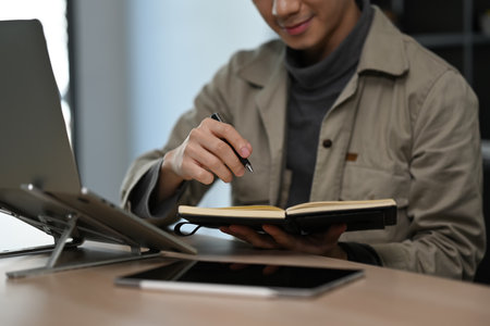 Cropped image of man freelancer working online with laptop and writing records to daily planner.