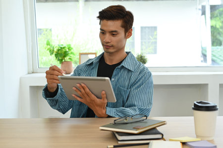 Handsome asian male freelancer checking online information on digital tablet, working on modern home office.の写真素材
