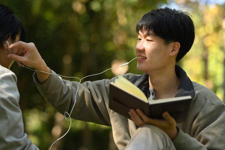 Image of young couple reading a book and listening to music in earphones while relaxing at public park together.の写真素材