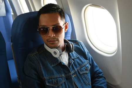 Hipster man passenger listening to music in wireless headphones while sitting in the airplane next...