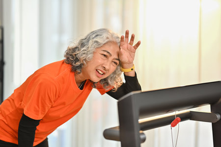 Tired senior woman wiping sweat of her forehead after rinning on a treadmill indoors. Healthy active...