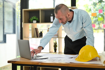Smart engineer man in shirt using laptop and working with blueprints, planning development details at workstation.の写真素材