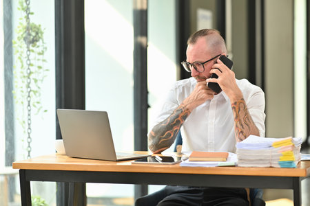 Handsome adult man employee in white shirt and glasses having phone conversation while sitting at desk in modern office.の写真素材