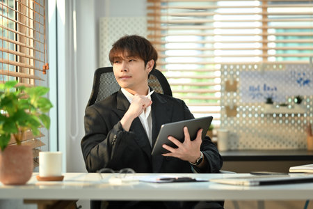 Thoughtful businessman holding digital tablet and looking through window, pondering idea or developing business strategy.の写真素材