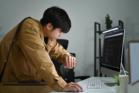 Busy male programmer standing in front of computer monitor with coded data and checking time on his watch.の写真素材