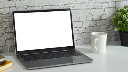 Creative workplace with laptop, coffee cup, potted plant and book on white table against brick wall.