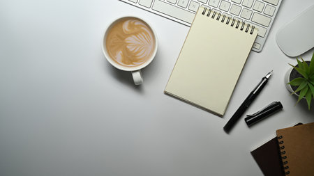 Top view of spiral notepad, pen, cup of coffee and keyboard on white office desk. Copy space for...