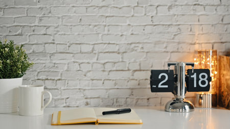 Home office desk with vintage flip clock and books and houseplant on white table against brick wall....