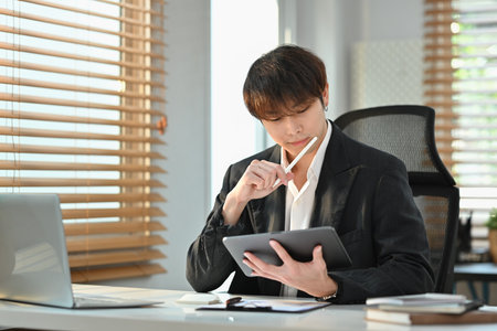 Image of financial analyst in elegant suit sitting at his workstation and analyzing statistical data on digital tablet.の写真素材
