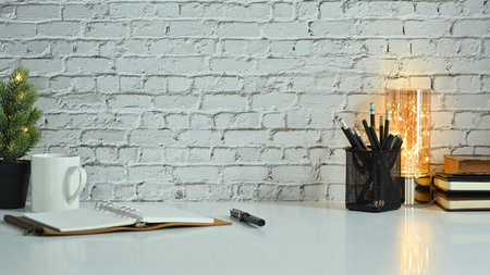 Notebook, potted plant, pencil holder and coffee cup on white table against brick wall. Stylish...