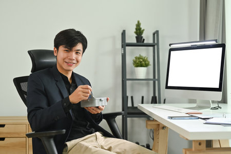 Smiling asian male worker sitting at office desk with blank computer screen, enjoying lunch break in office.の写真素材