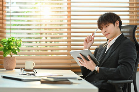 Portrait of asian male business leader sitting in comfortable workplace and reading email on digital tablet.の写真素材