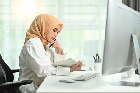 Focused millennial muslim businesswoman wearing hijab sitting at her workplace and checking working schedule plan.の写真素材