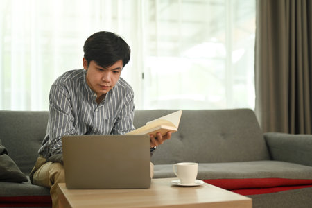 Relaxed asian man holding notebook and watching webinar or working online on laptop at home.の写真素材