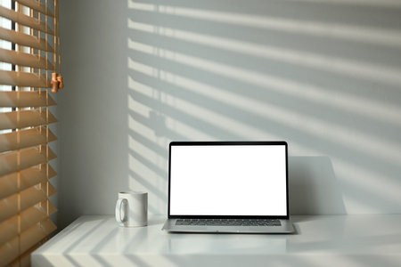 Front view of laptop computer and cup of coffee on white wooden table with sunlight from blinds window.の写真素材