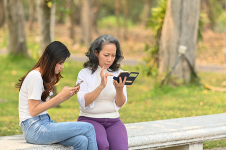 Image of middle age woman and daughter using mobile phone while sitting on a bench in calm nature park.の写真素材