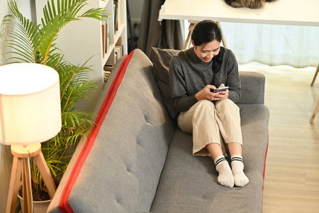 Overhead view of young woman relaxing on couch at cozy home browsing wireless internet or chatting online on smartphone.の写真素材