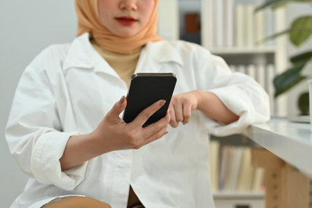 Smiling young Muslim woman employee in hijab sitting at her working desk and chatting online on...
