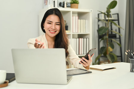 Cheerful asian woman worker holding mobile phone and communicating or online watching webinar on laptop.の写真素材