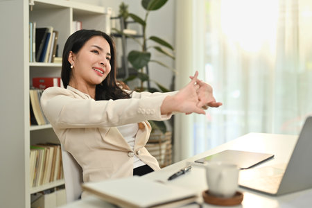 Pleasant young working woman in formal outfit stretching arms, relaxing on comfortable office chair.の写真素材